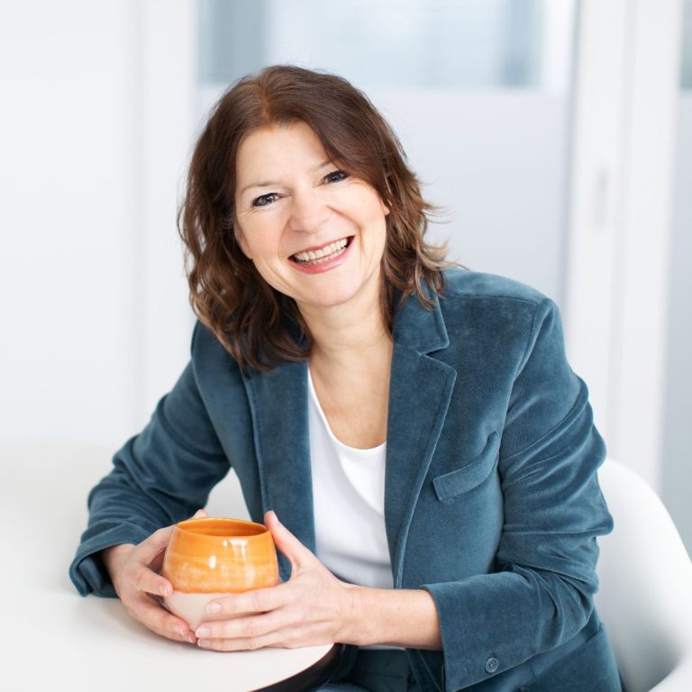 Frau mit lockigem, braunem Haar, die lächelnd eine orangefarbene Tasse hält.